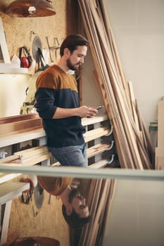 Designer craftsman looking casual and using his phone to send a message while standing in his studio workshop