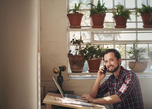 Male designer working at his studio desk while talking on the phone and looking up to smile at the camera