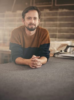 Casually dressed young man leaning on a work surface in his studio and looking away in a positive yet thoughtful manner
