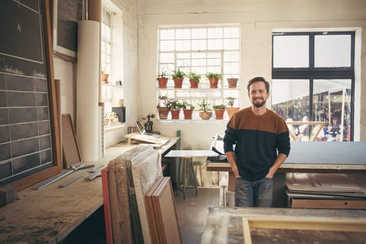 Young male craftsman standing with his hands in his pockets, in his workshop and smiling at the camera in a way that is both confident and comfortable
