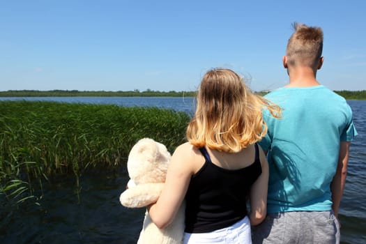 Two young individuals stand peacefully by a tranquil lake, happily embracing nature while holding a teddy bear