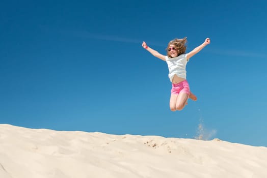 Young girl jumps from a dune in front of a blue sky