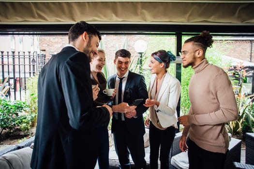 Small group of business people are standing in a cafe courtyard with hot drinks. They are discussing something on a smart phone.