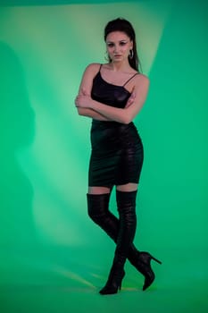 A stylish model poses confidently in a black dress and thigh-high boots on a vibrant green backdrop.