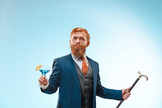 The bearded man in a suit holding cane and cocktail. Isolated on a blue background.
