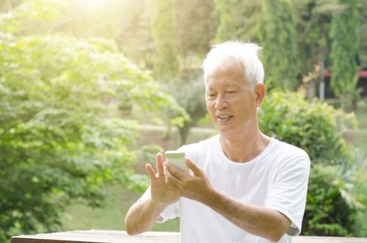 Portrait of cheerful gray hair Asian old man using smart phone, sitting at outdoor park in morning.