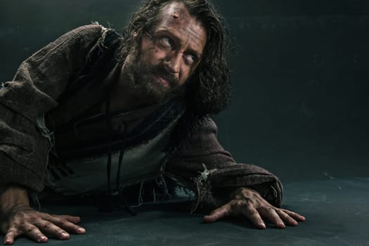Actor in makeup, a poor man on black studio background. The bum in canvas rags. Concept of vagrancy and survival. Concept of pain and suffering. The wounded man crawling to salvation