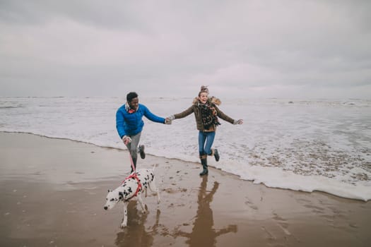 A young couple run away from the waves whilst walking their dog on a winter beach.