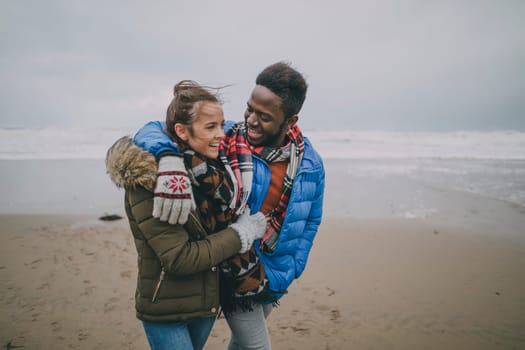 A young couple take a romantic stroll along a winter beach with their arms around each other.