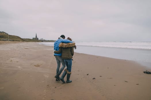 A young couple walk along a winter beach with their arms around each other.