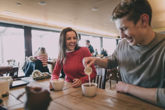 A young couple laugh in a cafe in winter whilst the male pours milk into a cup of coffee.
