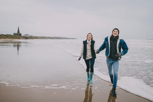 A young couple smile as they walk along a winter beach together.