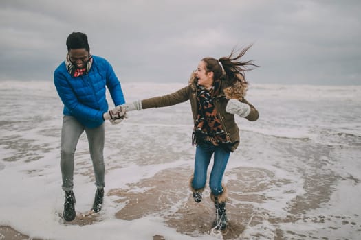 A young couple laugh, hold hands and paddle in the sea on Tynemouth beach in winter.