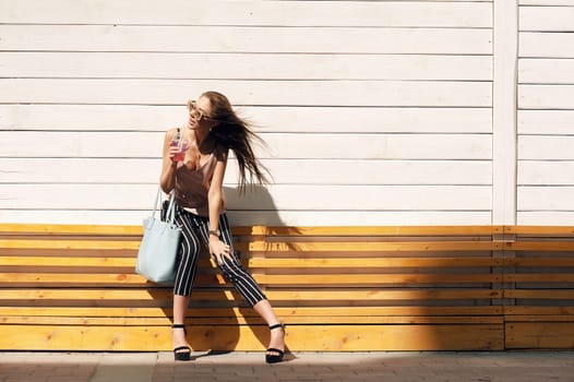 Outdoor portrait of a young beautiful fashionable happy lady wearing sunglasses, in a wooden frame, against a wooden, white wall. The model wears stylish clothes, a beige blouse, black breeches in a white stripe, a bag of light blue. The girl looks away and drinks pink mors and tubes. Women's fashion. Urban way of life.