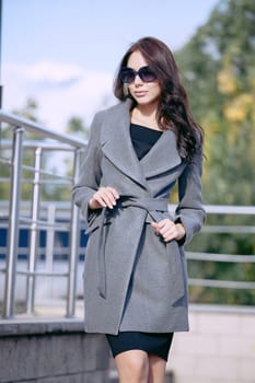 Outdoor portrait of a young beautiful fashionable woman, outdoors. A model dressed in a stylish gray coat, sunglasses. The concept of women's fashion, urban lifestyle.