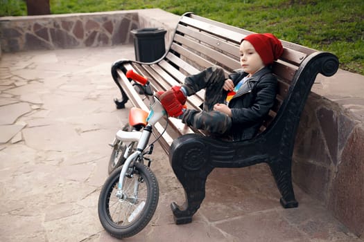 Portrait of a cute boy boy in a red cap-cap, sitting on a bench in a city park, with his bike and looking into the distance. Rest after a bicycle. Child outdoors. Emotion fatigue
