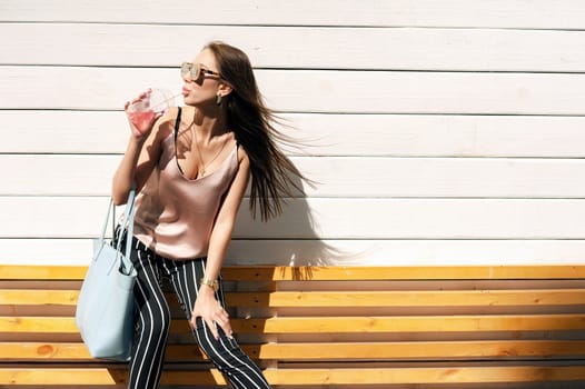 Outdoor portrait of a young beautiful fashionable happy lady wearing sunglasses, in a wooden frame, against a wooden, white wall. The model wears stylish clothes, a beige blouse, black breeches in a white stripe, a bag of light blue. The girl looks away and drinks pink mors and tubes. Women's fashion. Urban way of life.