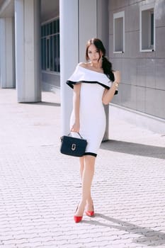 Young beautiful stylish girl in white dress and black bag. walk and pose outdoors. fashion style, shopping. The concept of women's fashion, urban lifestyle