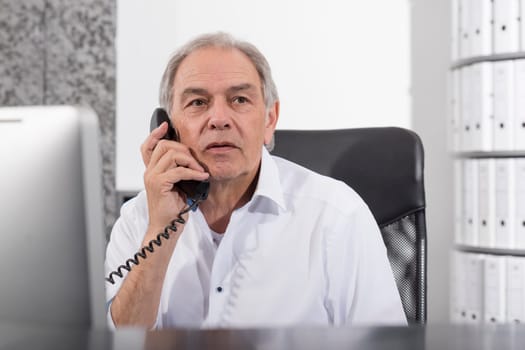 Elderly man on the phone with a white shirt sits at a desk