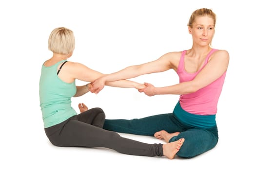 Side full-body view of two young blonde women sitting on the floor,touching each other on the soles of their feet and holding hands during a partner yoga exercise against a white background with slight shadows.