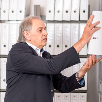 Elderly man in a suit leafs through in front of a shelf wall with documents in a folder