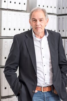 Older man in a suit stands in front of a shelf wall with documents folder