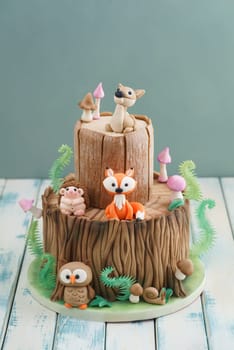 Enchanted forest woodland themed fondant cake with a hedgehog, deer, owl, fox, snail, tree trunk, ferns, mushrooms and leaves on wooden background