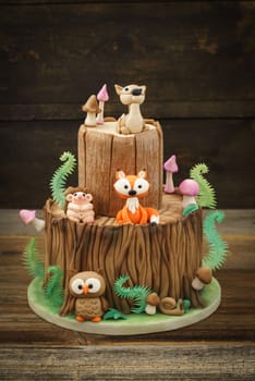 Enchanted forest woodland themed fondant cake with a hedgehog, deer, owl, fox, snail, tree trunk, ferns, mushrooms and leaves on wooden background