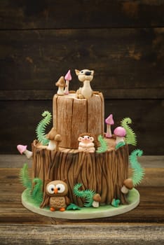 Enchanted forest woodland themed fondant cake with a hedgehog, deer, owl, tree trunk, ferns, mushrooms and leaves on wooden background