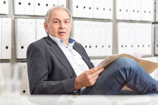 Older man in a suit sits in front of a shelf wall with documents folder