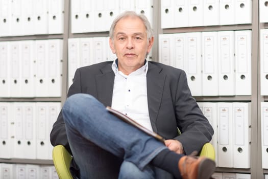 Older man in a suit sits in front of a shelf wall with documents folder