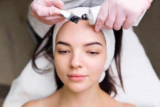 cosmetologist applying black mask on pretty woman face wearing black gloves, gorgeous woman in spa having facial procedures.