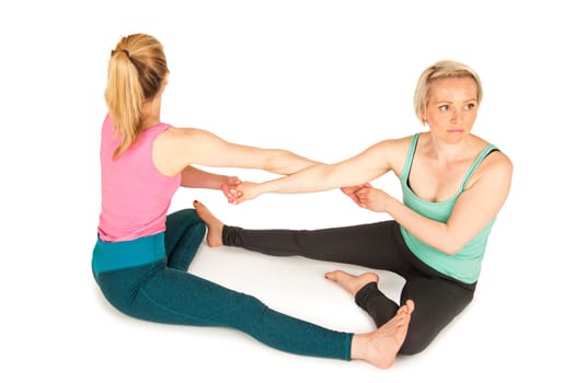 Elevated side full-body view of two young blonde women sitting on the floor,touching each other on the soles of their feet and holding hands during a partner yoga exercise against a white background with light shadows.