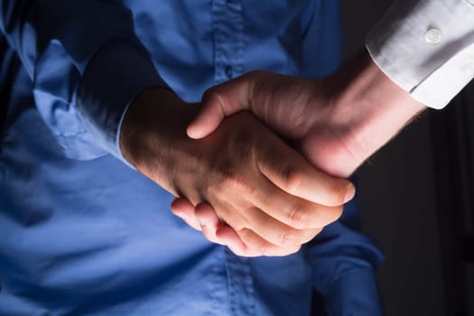 Low angle view of two businessmen hands shaking over dark gray background and light source at bottom side