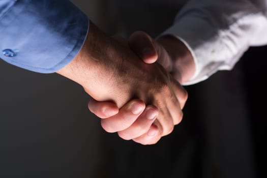 Low angle view of two businessmen hands shaking over dark gray background and light source at bottom side