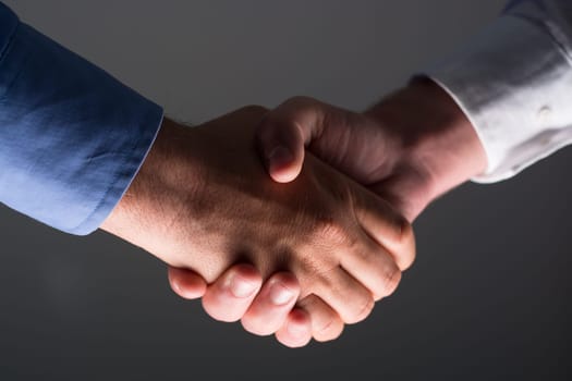 Low angle view of two businessmen hands shaking over dark gray background and light source at bottom side