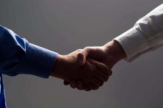 Low angle view of two businessmen hands shaking over dark gray background and light source at bottom side