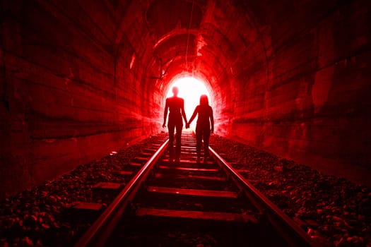 Couple walking hand in hand along the track through a railway tunnel towards the bright light at the other end, they appear as silhouettes against the light