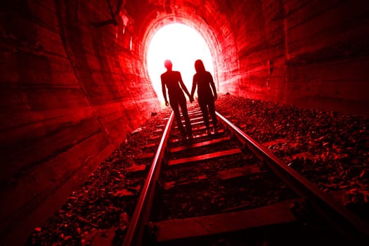 Couple walking hand in hand along the track through a railway tunnel towards the bright light at the other end, they appear as silhouettes against the light