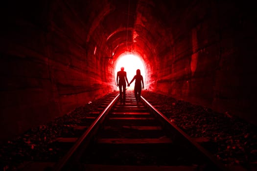 Couple walking hand in hand along the track through a railway tunnel towards the bright light at the other end, they appear as silhouettes against the light