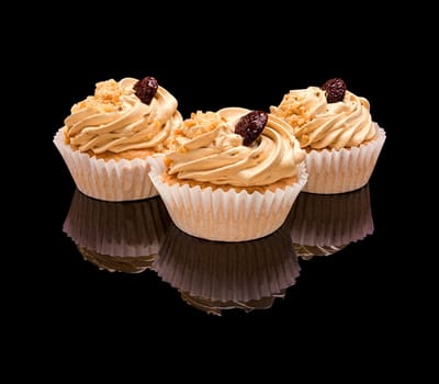 Three muffin cupcake with coffee cream, chocolate and pieces of nuts in a paper cup. On a black glossy background with real reflection. Isolated on black