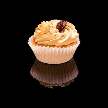 One muffin cupcake with coffee cream, chocolate and pieces of nuts in a paper cup. On a black glossy background with real reflection. Isolated on black
