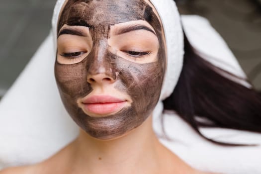 cosmetologist applying black mask on pretty woman face wearing black gloves, gorgeous woman in spa having facial procedures.