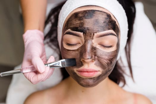 cosmetologist applying black mask on pretty woman face wearing black gloves, gorgeous woman in spa having facial procedures.