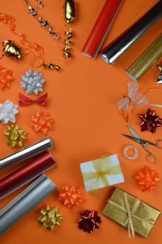 gift wrapping utensils on orange ground in autumn colors red, orange, golden, silver and white with rolls of wrapping paper, gift ribbons, bow topper, scissors and cellotape