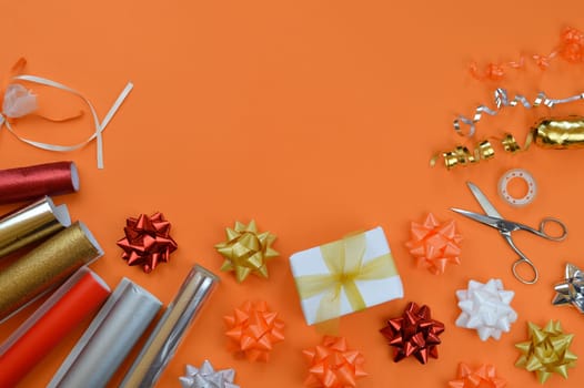 gift wrapping utensils on orange ground in autumn colors red, orange, golden, silver and white with rolls of wrapping paper, gift ribbons, bow topper, scissors and cellotape
