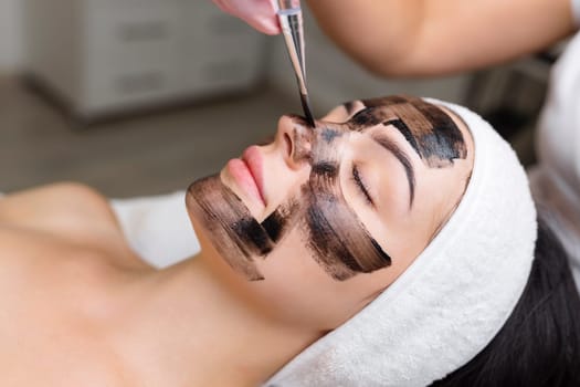 cosmetologist applying black mask on pretty woman face wearing black gloves, gorgeous woman in spa having facial procedures.