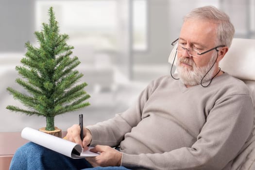 Old man making a shopping list with a pen on paper next to an undecorated fir tree for Christmas holidays