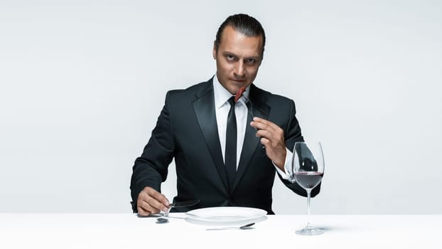 Portrait of a young man in image of maniak with knife, fork and and a piece of raw meat on a plate. Man in suit. The image in the style of Halloween