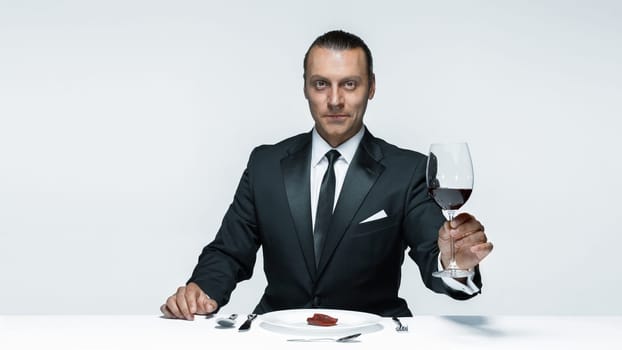 Portrait of a young man in image of maniak with knife, fork and and a piece of raw meat on a plate. Man in suit. The image in the style of Halloween